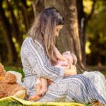 Tender moment of a mother breastfeeding her baby outdoors with a teddy bear nearby.