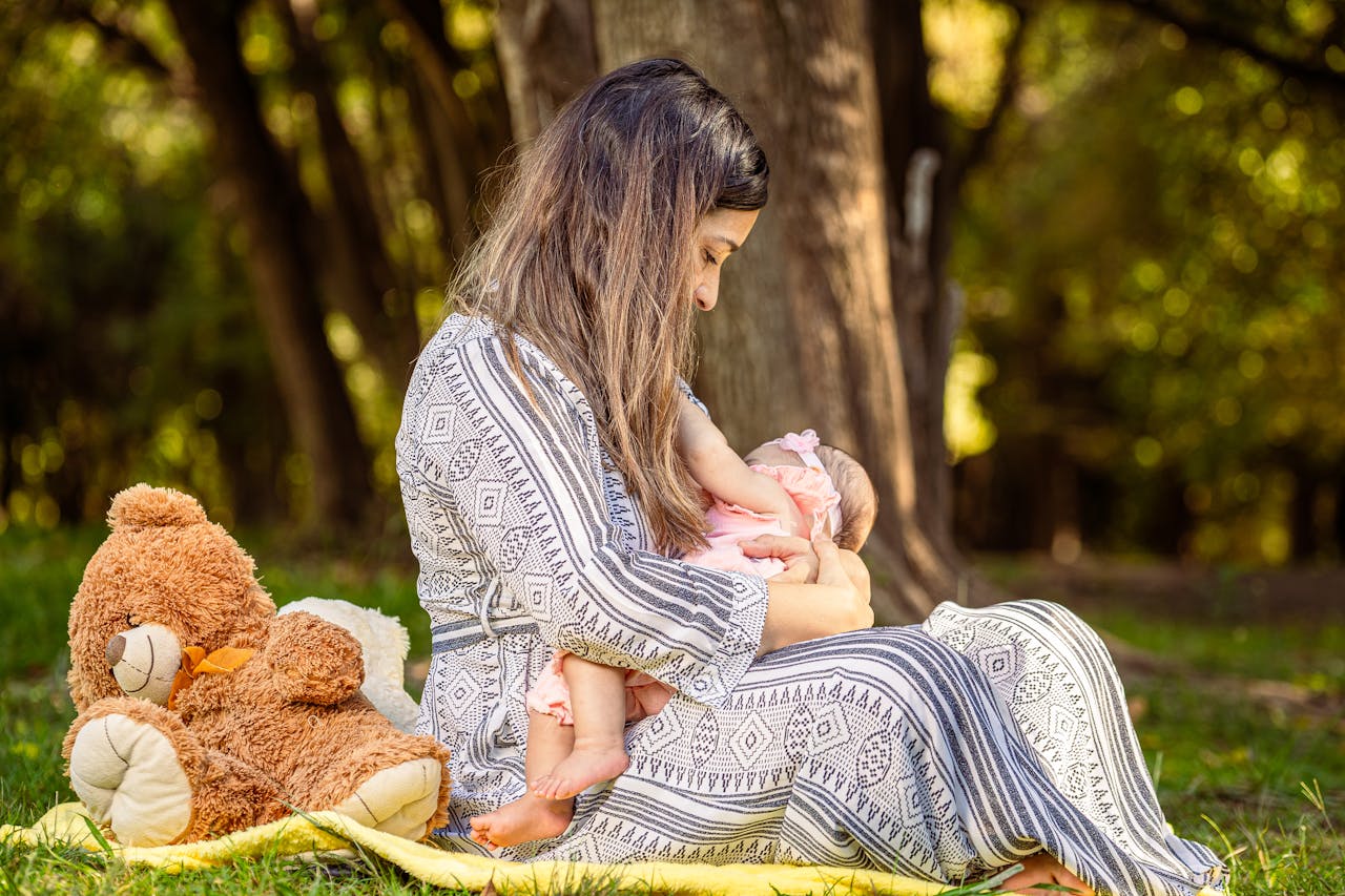 Tender moment of a mother breastfeeding her baby outdoors with a teddy bear nearby.